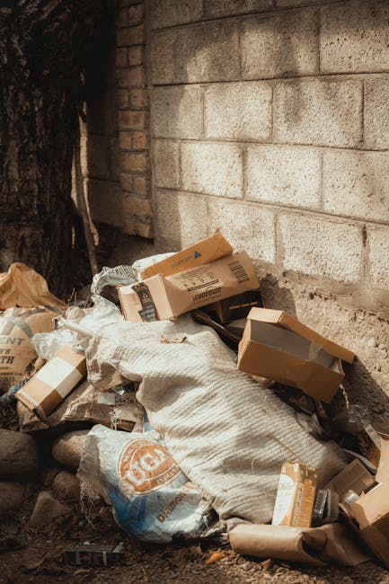 A pile of mixed waste consisting of flattened cardboard boxes, some partially opened and others intact, along with a large, worn white fabric sack, is situated on the ground near a brick and concrete wall. The boxes are light brown with visible folds and creases, and some feature printed labels or barcodes. The material of the sack appears rough and coarse, likely woven, with areas showing dirt or discoloration. The waste is positioned at the base of the wall, close to a dark tree trunk on the left side of the image, which partially obscures the corner. The background includes a weathered brick wall topped with a concrete block wall, both displaying signs of age and exposure without fresh paint or finishing. The overall scene suggests an outdoor environment where private rubbish collection or on-site clearance might be occurring, aligning with services provided by companies like Rubbish Removal Enfield for alternative waste handling options.