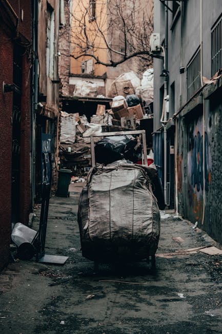 A pile of mixed waste consisting of flattened cardboard boxes, some partially opened and others intact, along with a large, worn white fabric sack, is situated on the ground near a brick and concrete wall. The boxes are light brown with visible folds and creases, and some feature printed labels or barcodes. The material of the sack appears rough and coarse, likely woven, with areas showing dirt or discoloration. The waste is positioned at the base of the wall, close to a dark tree trunk on the left side of the image, which partially obscures the corner. The background includes a weathered brick wall topped with a concrete block wall, both displaying signs of age and exposure without fresh paint or finishing. The overall scene suggests an outdoor environment where private rubbish collection or on-site clearance might be occurring, aligning with services provided by companies like Rubbish Removal Enfield for alternative waste handling options.