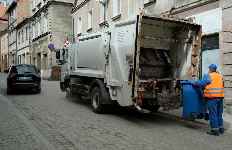 A white stationary rubbish collection truck with an open rear compartment positioned on a narrow urban street paved with cobblestones. The truck's rear is designed for waste collection, showing rusted metal and mechanical components exposed, indicating it is actively loading or ready to load waste. To the right, a waste collection worker wearing a blue uniform, orange high-visibility vest, and matching blue gloves is positioned beside a blue wheelie bin, guiding or preparing it for collection. Behind the worker and truck, the surrounding environment includes a row of old, multi-story buildings with varying textures and muted colors, such as beige and light grey, with some weathered facades. A black car is parked on the left side of the image, closer to the curb, and partial street signs are visible attached to one building, signaling parking restrictions. The lighting is neutral, suggesting overcast conditions, and the scene depicts an organized waste collection process in an urban residential area, emphasizing private or independent rubbish removal services consistent with the context of rubbish management in Enfield.