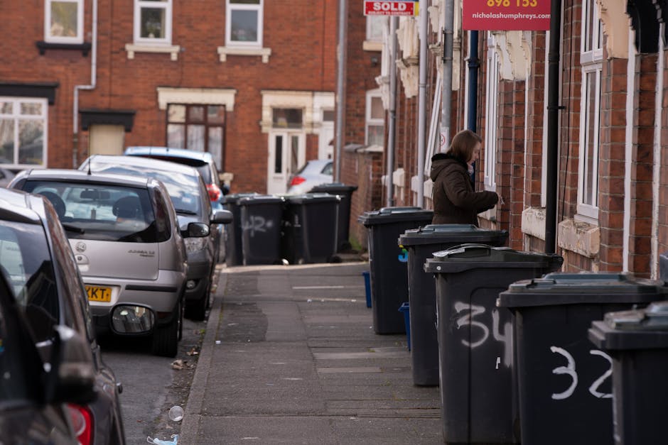 A residential street scene shows several black wheelie bins lined up along the sidewalk outside brick terraced houses. The bins are made of plastic with textured surfaces and are fitted with gray lids, some of which are slightly ajar. The sidewalk is paved with concrete slabs, exhibiting a weathered surface with minor cracks and some scattered debris. In the background, parked cars of various models and colors, including silver and black, occupy roadside parking spaces adjacent to the bins. A woman dressed in a dark jacket stands beside one of the bins, appearing to be preparing for rubbish disposal or collection. The brick facades of the houses feature white-framed windows with small decorative stone sills, and one house has a sign referencing private or independent waste collection services. Overhead, the scene is under natural daylight, with soft shadows cast on the street, suggesting an overcast or partly cloudy day. This image exemplifies typical urban rubbish collection practices, likely coordinated by private waste management companies such as Rubbish Removal Enfield, serving local residential areas with on-site waste disposal options outside homes.