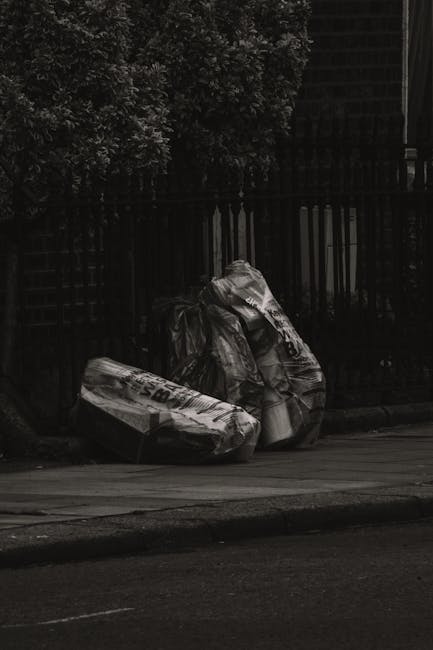 A pile of discarded packaging materials and rubbish placed against a concrete wall in an outdoor setting. The waste includes flattened cardboard boxes, some partially crushed or torn, and a large white sack containing additional debris. The cardboard items vary in shades of brown, with some featuring printed labels and barcodes, while others are plain. The pile is situated on a bed of small rocks and dirt, adjacent to a brick wall structure visible in the background, along with a tree trunk on the left side of the image. The overall scene conveys an uncollected waste accumulation typical of private disposal or on-site clearance, aligning with services offered by Rubbish Removal Enfield to address such rubbish buildup efficiently. Natural light illuminates the scene, highlighting the textures of the cardboard surfaces and the roughness of the surrounding environment.