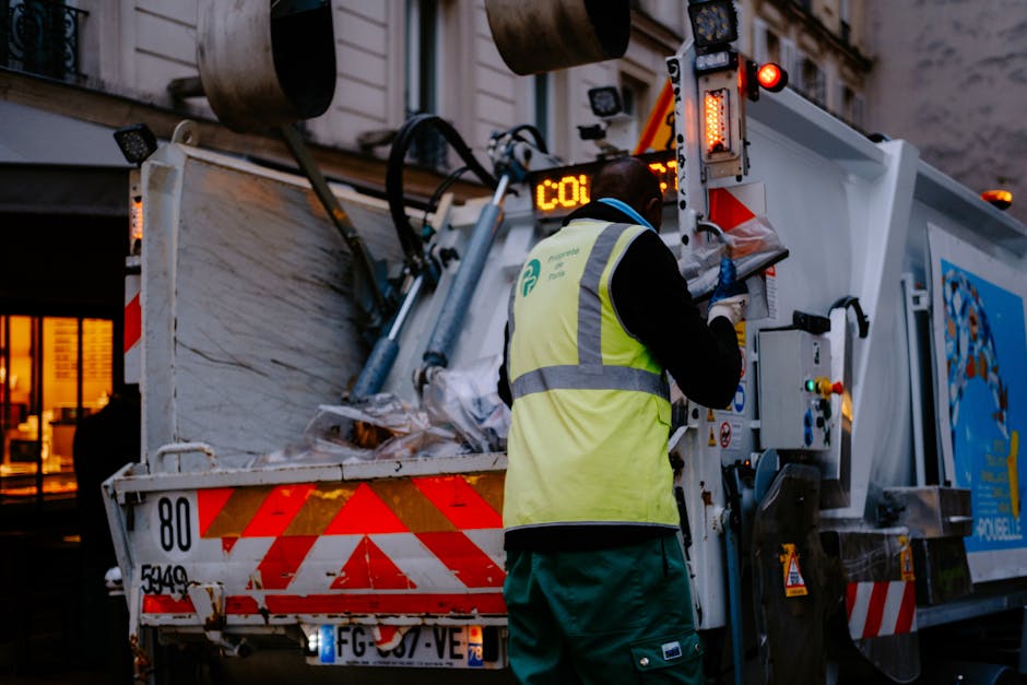 A worker wearing a high-visibility yellow vest and dark clothing is operating a waste collection vehicle on a city street during the evening. The back of the vehicle is open, revealing a load of mixed waste, including paper and plastic materials, with a textured, metallic surface that appears slightly weathered. The vehicle features reflectors with red and white chevron patterns, and various hydraulic components and control panels are visible on the side where the worker is positioned. In the background, there are multi-story buildings with lit windows, indicating a populated urban environment. The scene is illuminated by artificial lighting, casting a warm glow, and the worker appears to be engaged in manual control of the vehicle's operations, supporting private or independent waste collection services that handle rubbish removal outside regular municipal systems, consistent with services offered by Rubbish Removal Enfield.