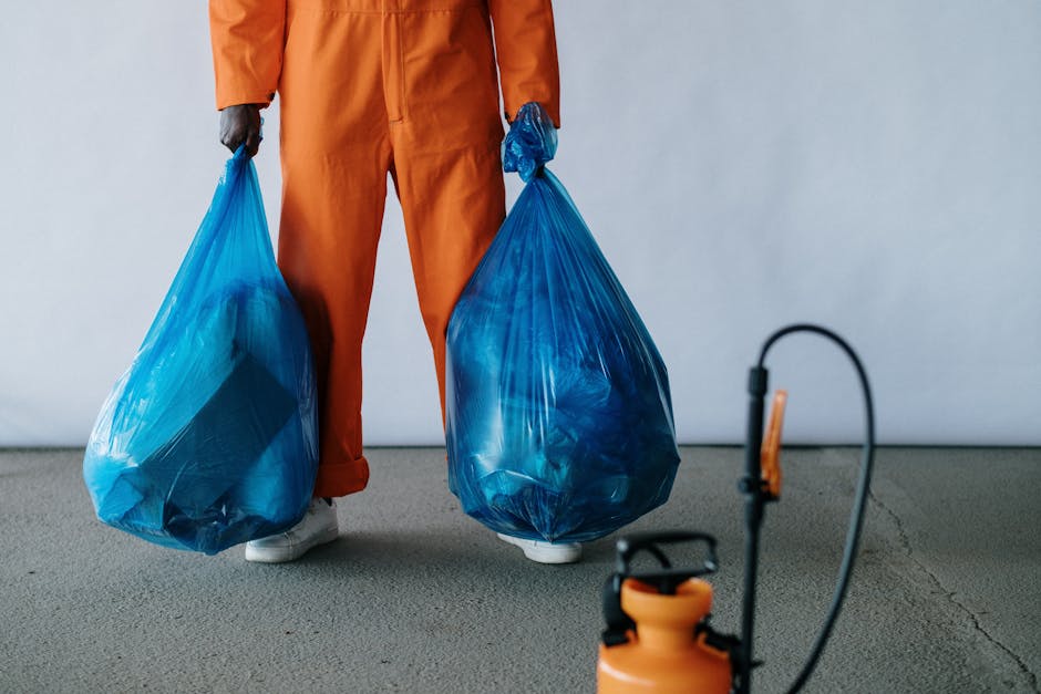 A person dressed in an orange work jumpsuit and white safety shoes is standing on a smooth concrete floor, holding two large blue plastic garbage bags filled with waste, one in each hand. The bags are semi-transparent, revealing their contents, and are tied at the top. The background is a plain, light grey wall, and part of a black and orange device with a flexible hose and a small handle is visible in the foreground on the right side. This scene depicts a waste collection or rubbish removal activity that could be associated with private or independent disposal services, such as those offered by Rubbish Removal Enfield, and highlights the physical process involved in on-site rubbish clearance.