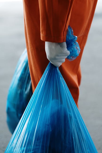 A worker wearing a high-visibility yellow vest and dark clothing is operating a waste collection vehicle on a city street during the evening. The back of the vehicle is open, revealing a load of mixed waste, including paper and plastic materials, with a textured, metallic surface that appears slightly weathered. The vehicle features reflectors with red and white chevron patterns, and various hydraulic components and control panels are visible on the side where the worker is positioned. In the background, there are multi-story buildings with lit windows, indicating a populated urban environment. The scene is illuminated by artificial lighting, casting a warm glow, and the worker appears to be engaged in manual control of the vehicle's operations, supporting private or independent waste collection services that handle rubbish removal outside regular municipal systems, consistent with services offered by Rubbish Removal Enfield.